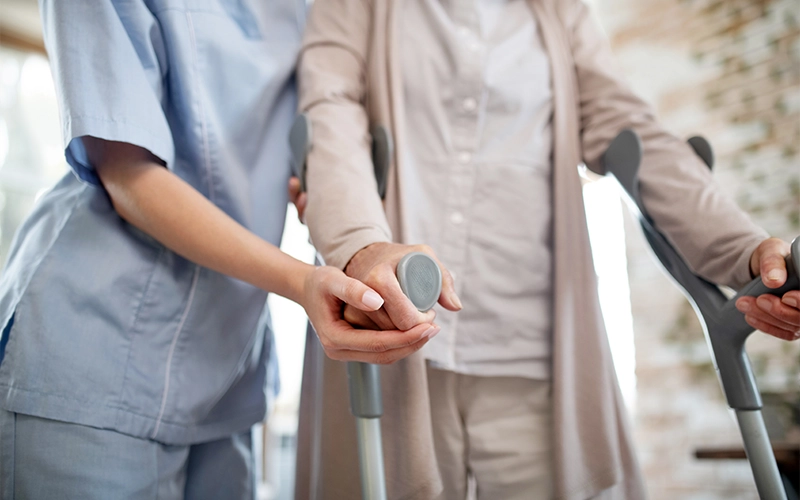 מטפלת עוזרת למטופלת בקביים ללכת, מחבקת אותה בשני צדדים