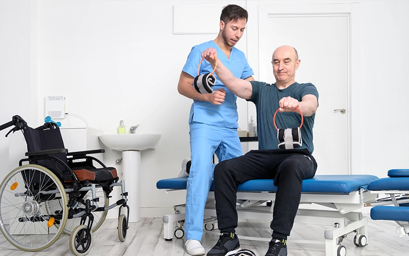 פעילות גופנית, מטופל יושב על המיטב מחזיק משקולות בשתי ידיים ומטפל מוסמך עומד לידו עוזר ומשגיח עליו